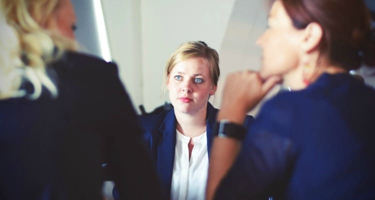 Three women having a discussion in an office setting, with the woman in the center looking directly ahead and the other two women looking at her.