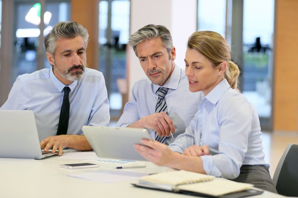 Three professionals in a meeting, two men and a woman, are looking at a tablet. They are seated at a table with laptops, documents, and a notebook in a modern office setting.