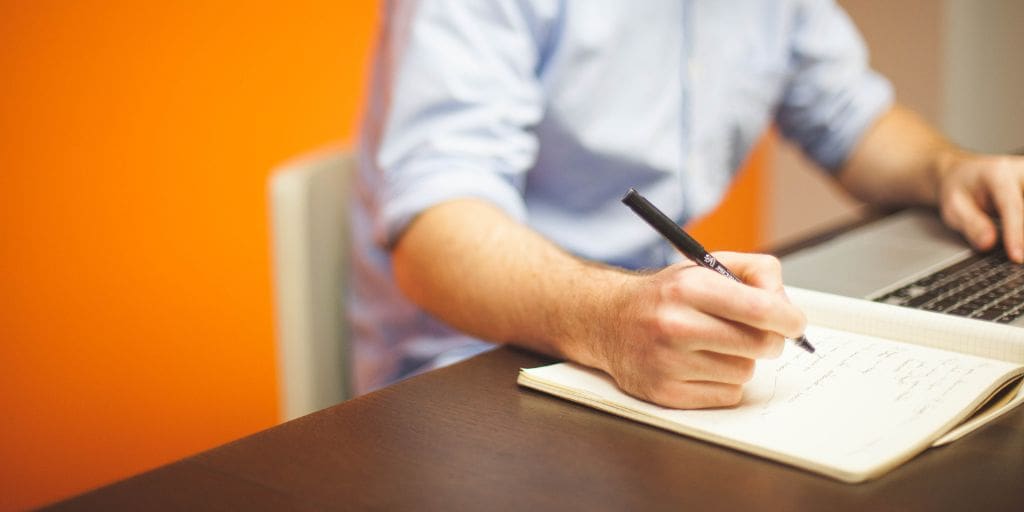 A person in a light blue shirt writes in a notebook with a pen while sitting at a desk next to an open laptop. The background features an orange wall.