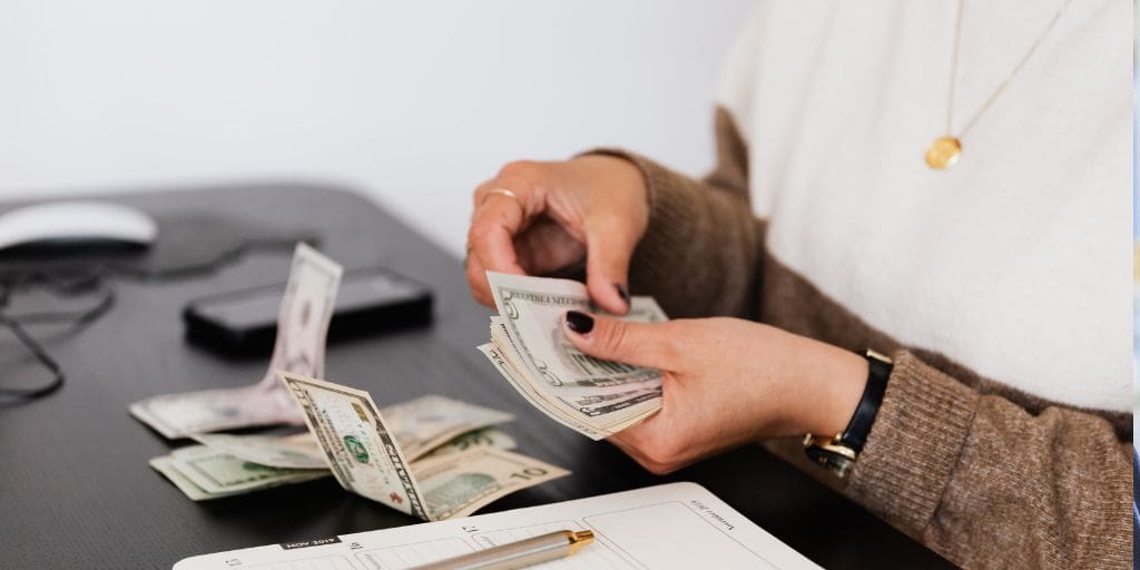 Person counting a stack of U.S. banknotes at a desk, with scattered bills, a pen, and a paper on the surface.