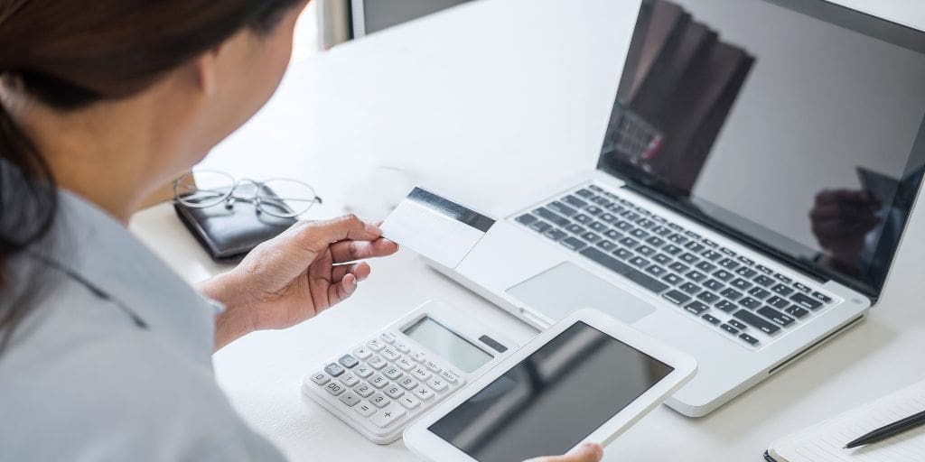 Person holding a credit card and a tablet in front of a laptop, with a calculator and eyeglasses nearby on the desk.