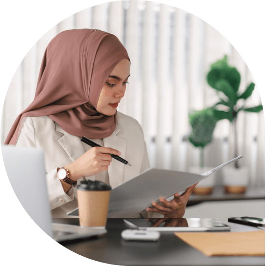 A person wearing a hijab sits at a desk reviewing documents, holding a pen, with a coffee cup and laptop nearby.