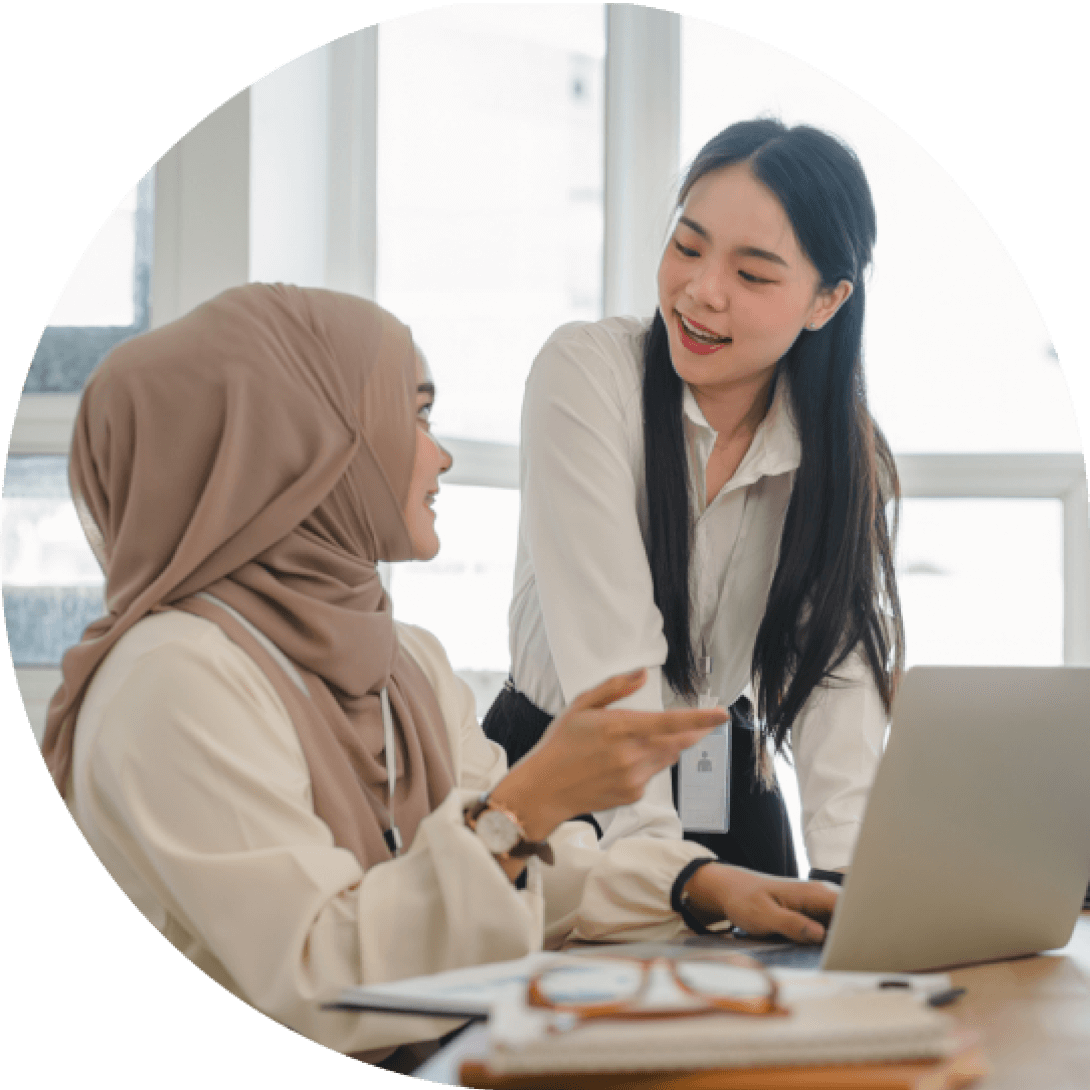 Two women at a desk, one wearing a hijab, are engaged in conversation with a laptop and documents in front of them.